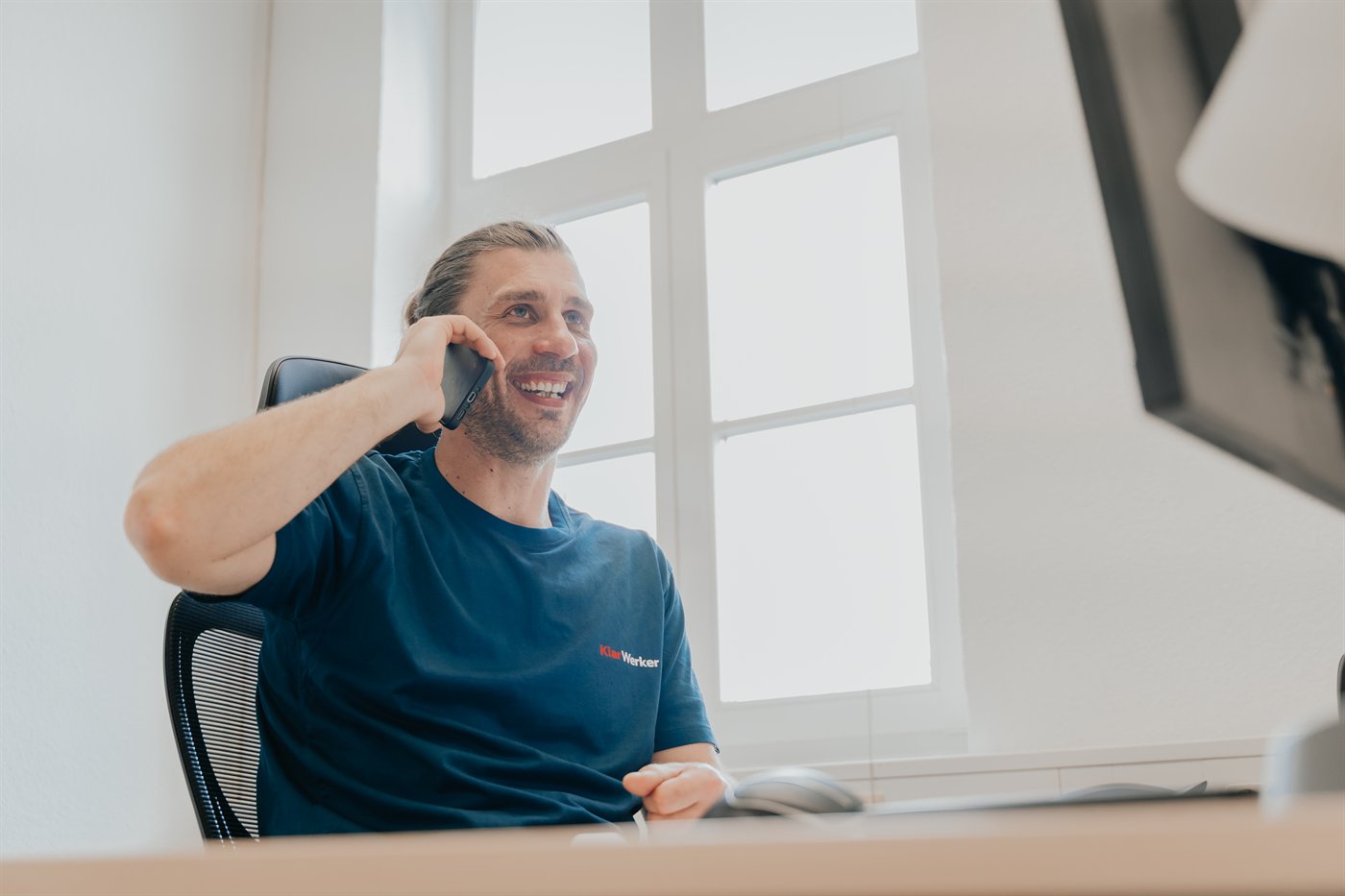 Marco Schulz von KlarWerker lacht beim Telefonat am Schreibtisch, im KlarWerker-Shirt